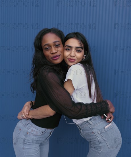 Two young women embrace in a joyful, friendly hug against a vibrant blue background. Their radiant smiles and casual attire create a warm, inviting atmosphere of friendship