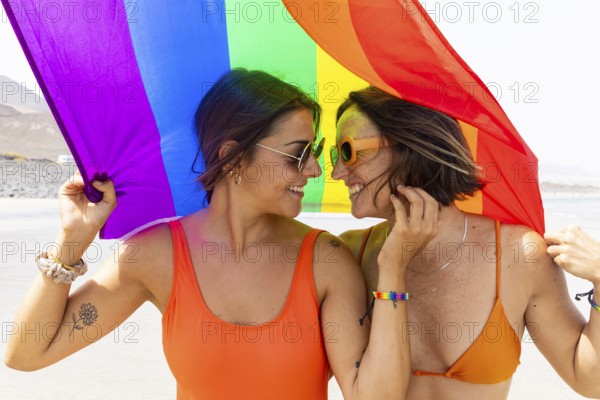 A joyful lesbian couple shares a moment under a colorful rainbow flag on a sunny beach. Both are wearing matching orange swimsuits and sunglasses, symbolizing love and diversity