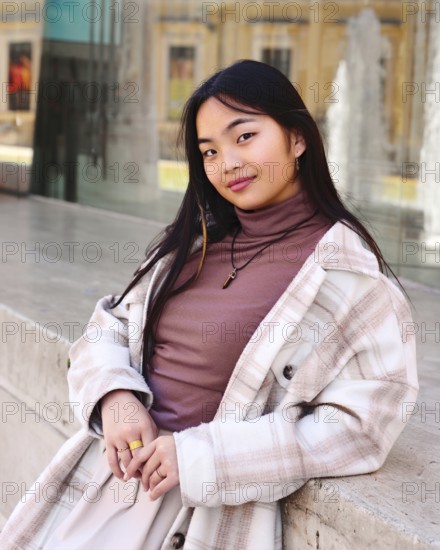 A young chinese woman with long dark hair wearing a plaid coat and brown turtleneck leans casually against a stone surface, with a modern water fountain in the background