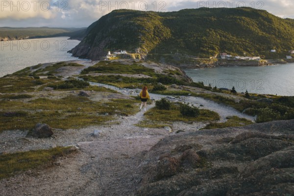 Back view of an unrecognizable person walking along a rugged trail with a panoramic vista of Signal Hill, St. John's. The person is wearing a backpack and exploring the scenic coastal landscape during sunset
