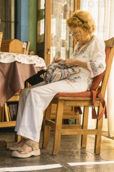 Elderly woman wearing glasses and white clothes is focused on sewing a colorful fabric while comfortably sitting on a wooden chair with a red cushion