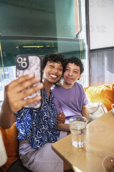 A joyful, multiethnic LGBTQ+ couple sits in a cozy cafe, smiling as they take a selfie. Sunlight streams through the window, illuminating their happy expressions