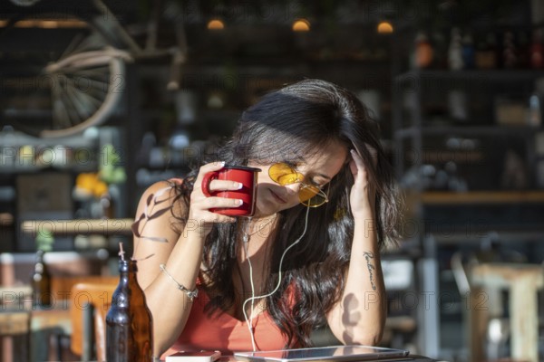 Woman in a cafe, savoring coffee with yellow sunglasses and earphones, enjoying music on a sunny day. The ambiance is relaxed and inviting, with a rustic decor backdrop