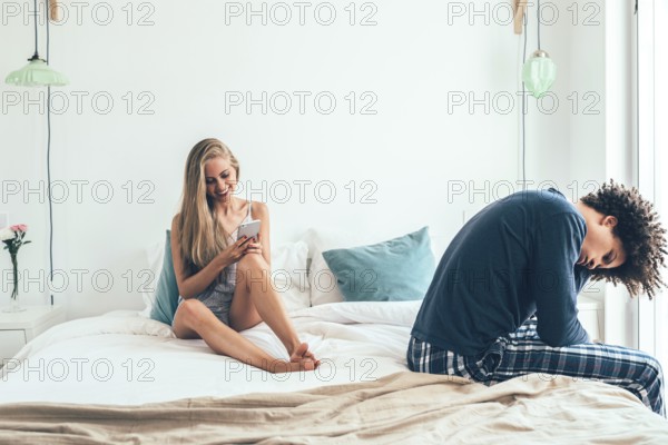 A woman smiles while using her phone on a bed, while a man sits with a sad expression, suggesting a moment of disconnect or relationship tension