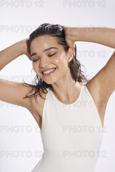 A young female model with wet hair and scattered drops of water on her face is captured smiling joyously while touching her head, dressed in a sleek white bodysuit against a plain background in a photo studio