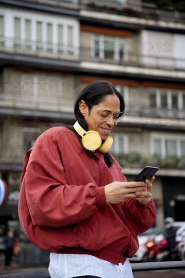 A man with long hair and large glasses wearing a red jacket and yellow headphones is outside looking at a smartphone, with an urban building in the background