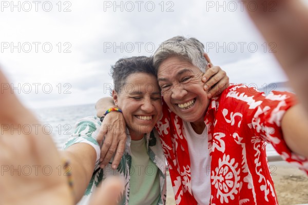 A joyful lesbian couple takes a selfie on a cloudy beach day, showcasing their love and excitement for their upcoming summer holidays