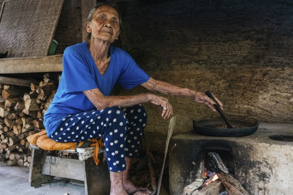 An elderly woman in a rustic kitchen prepares balinese coffee on a traditional wood fired stove. Dressed in a blue shirt and polka dot pants, she encapsulates cultural heritage and simple living