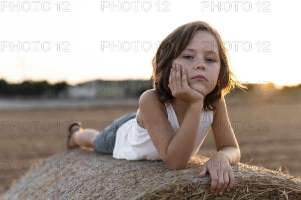 Child in casual attire lies on a hay bale during sunset, reflecting a calm and serene mood. The warm sunlight enhances the peaceful rural atmosphere