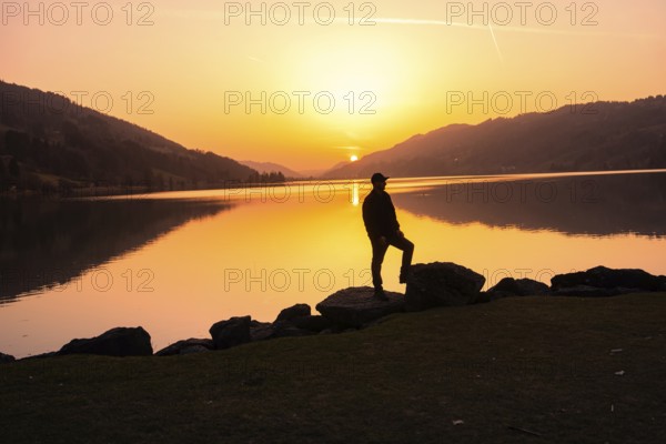 A person standing on rocks with a view of a calm lake at sunset, Großer Alpsee, Immenstadt im Allgäu, Bavaria, Germany