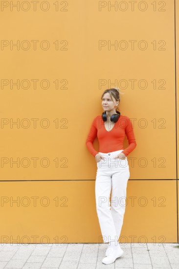 A fashionable woman leans against a striking orange wall in Warsaw, wearing bright attire with wireless headphones around her neck, exuding urban style and connectivity