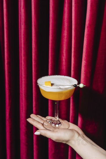 A peach cocktail served in an elegant coupe glass, delicately held against a backdrop of rich red curtains. Garnished with a candied peach and a pearl pick, this drink exudes luxury and refinement