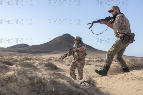 Two soldiers in camouflage gear patrol a sandy desert holding rifles. They observe their surroundings attentively under a clear blue sky with mountains in the background