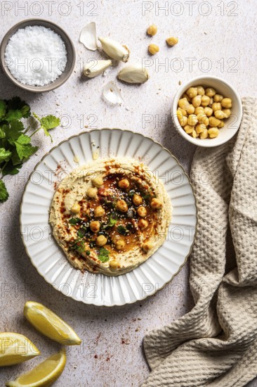 A beautifully styled hummus plate garnished with chickpeas, garlic, herbs, and spices on a textured surface, accompanied by lemon wedges and a cloth napkin