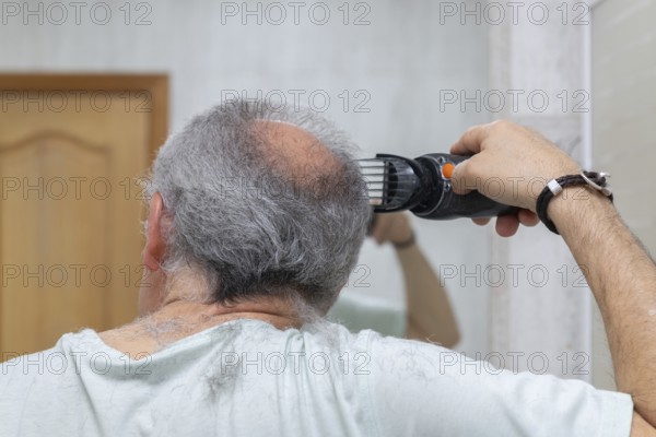 A man cuts his gray hair with electric clippers. The scene captures a home-style haircut in a casual, homey setting