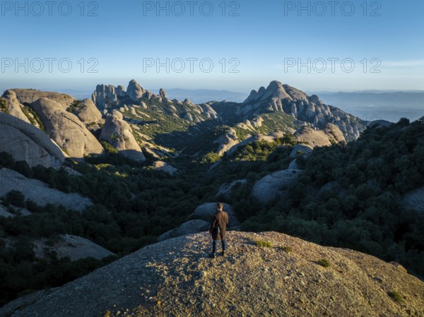 A person stands on a rocky hill, overlooking the stunning Montserrat mountains in Catalonia, Spain, with clear blue skies creating a breathtaking panorama