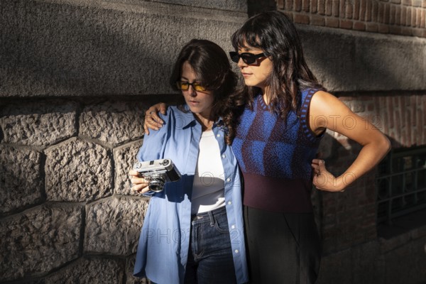 A multiethnic lesbian couple enjoys a moment together outdoors. One embracing the other while they look at a vintage camera. They are dressed stylishly, bathed in warm sunlight against a stone and brick wall