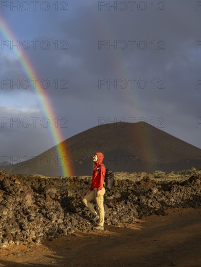 A woman in a red jacket stands on volcanic terrain in Timanfaya National Park, Lanzarote. A vivid rainbow arches across the sky, highlighting the contrasting natural beauty