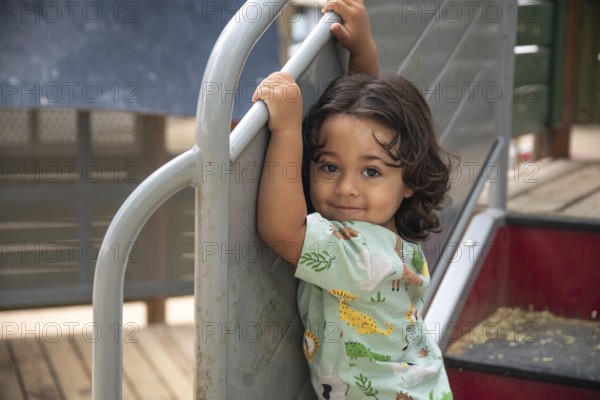 An happy toddler smiling and enjoying a playful day at the park. The child grips the playground equipment, reveling in outdoor fun and adventure during a sunny summer day