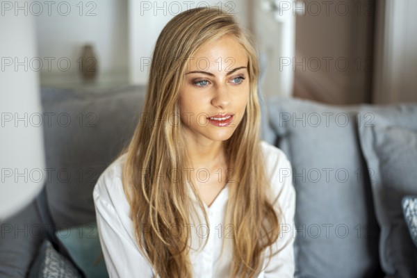A serene woman with long blonde hair sits on a comfortable gray sofa, looking thoughtfully into the distance. The cozy room offers a calm and inviting atmosphere