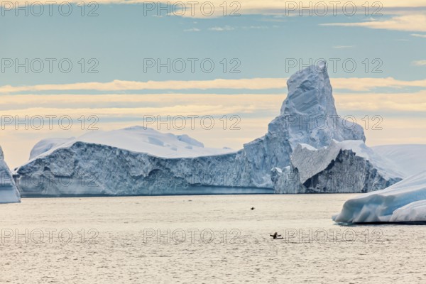 A large iceberg protrudes from the sea, a kayaker small below. The sky is cloudy, icebergs in the Southern Ocean in Antarctica