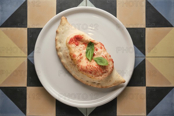 Freshly baked calzone garnished with basil leaves, served on a white plate over a vibrant geometric tiled surface
