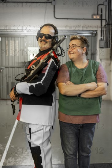 A blind shooting champion stands confidently with a supportive partner at an indoor shooting range. The champion holds a rifle, ready for competition or training