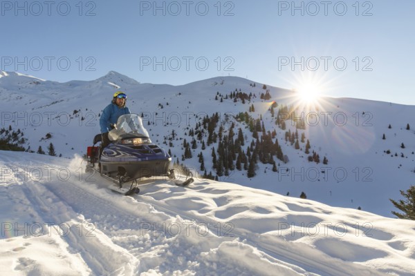 A snowmobiler rides through the picturesque snowy mountains, enjoying the stunning winter landscape with bright sunshine and snow-laden trees on a clear day