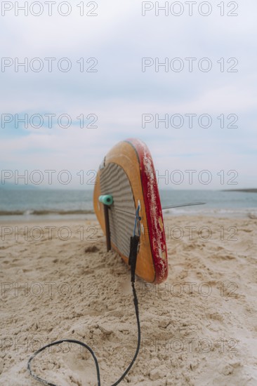 A surfboard lies on the sandy shore of a Peruvian beach, tethered by its leash, under a cloudy sky. The calm ocean extends into the horizon, inviting adventure
