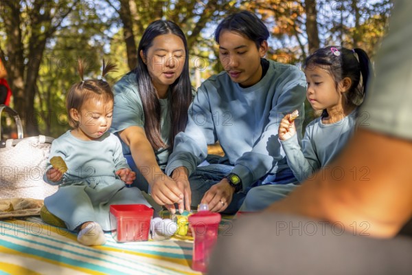 Asian family sharing a joyful picnic in a sunlit autumn park. They are seated on a blanket surrounded by colorful leaves, enjoying each other's company and a variety of snacks