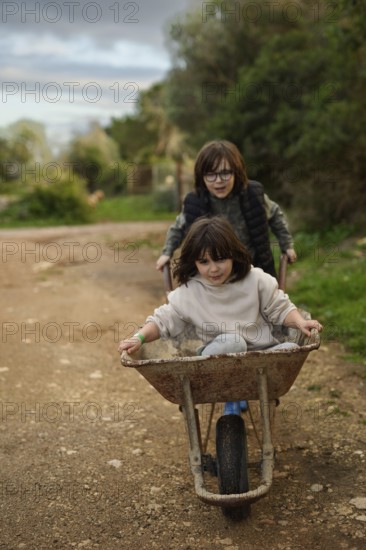 Two children enjoying a playful moment with a wheelbarrow on a rural path. The older child pushes while the younger one rides, surrounded by lush greenery and open space