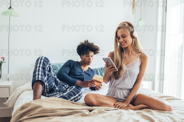 A couple enjoys a relaxed morning on a cozy bed, one listens to music on headphones, the other reads a book. The ambiance is calm and leisurely, perfect for unwinding