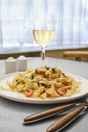 A plate of seafood pasta with shrimp and clams on a kitchen table. A glass of white wine stands nearby, complementing the meal in a bright and inviting dining space during midday