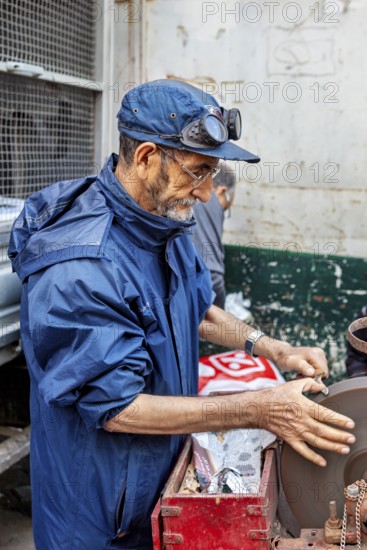 A man working on a machine in a workshop, wearing a blue jacket and a cap, A man sharpening knives in the historic centre of Algiers Algeria