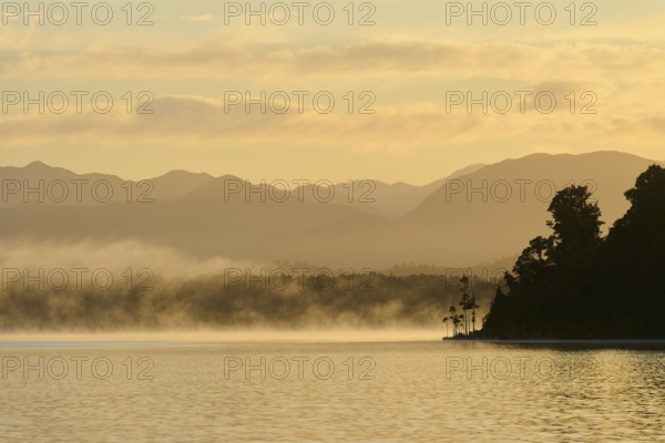 Mist drifts over a calm lake with mountains in the background at sunrise, Lake Mahinapua, Ruatapu, South Island, New Zealand