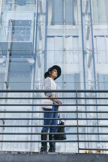 Stylish woman in a hat and fur-trimmed sweater stands by a modern glass building, exuding confidence and elegance. She holds a black bag, epitomizing urban fashion and modernity