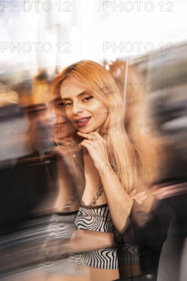 Trendy woman with long blonde hair poses confidently against a dynamic, blurred cityscape backdrop. The artistic motion blur adds a sense of urban energy and style