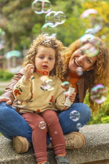 Vertical portrait of a cute blonde girl and mother sitting on a park blowing bubbles