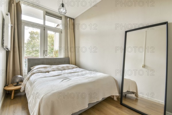 A stylish, serene bedroom featuring a bed with soft bedding, a large mirror, and bright natural light through large windows, creating a warm ambiance