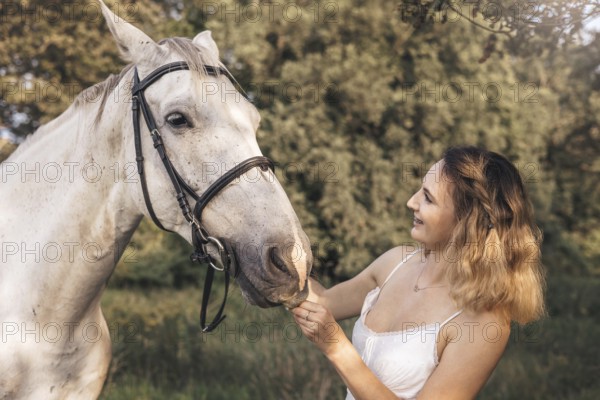 A young woman gently interacts with a white horse in a tranquil outdoor countryside environment. Sunlight filters through the trees, highlighting the connection between human and animal