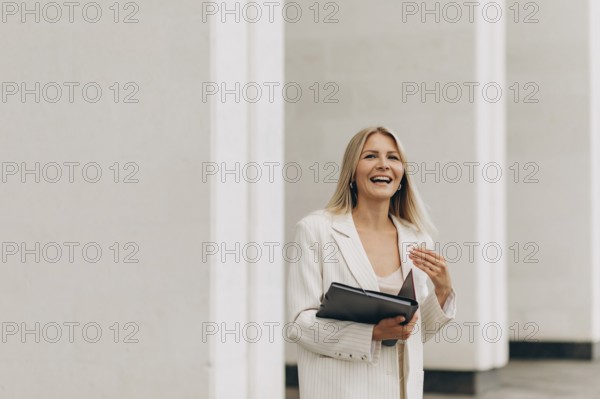 A businesswoman in a white suit smiles joyfully while holding a folder. She stands in front of sleek architecture, capturing a professional and optimistic demeanor