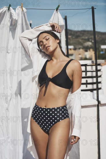 A young woman in a black polka-dot bikini and white shirt enjoys the summer sun on a rooftop The bright, airy setting creates a relaxed and fashionable vibe