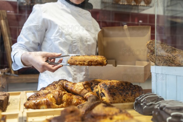 Bakery worker in uniform uses tongs to place a warm pain au chocolat into a paper takeaway box amid a tempting display of fresh pastries and bread on the counter