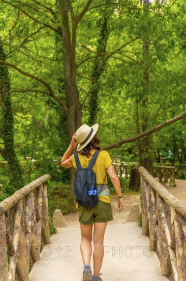 A woman wearing a straw hat and a green backpack is walking across a bridge. The bridge is surrounded by trees, and the woman is enjoying her walk
