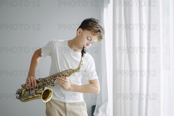 A young boy plays the saxophone in natural light by a large window. The casual outfit and serene setting convey a relaxed and creative atmosphere