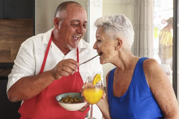A joyful senior couple engages in a playful and affectionate interaction in a bright kitchen during a party, sharing olives and orange juice