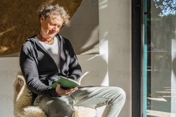 A man enjoys a quiet moment reading a book at home, sitting comfortably by a window, illuminated by warm sunlight The scene captures relaxation and peace