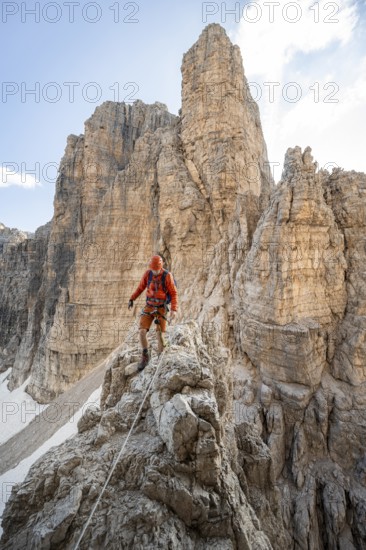 Mountaineer climbs on an exposed rock in the secured Via Ferrata Bocciere Centrale via ferrata, mountain landscape with spectacular rock towers in the back, Brenta-Adamello Nature Park, Trentino, Italy