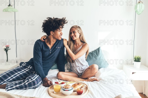 A young couple sits cheerfully on a bed, sharing a breakfast tray filled with fruit, juice, and pastries. The room is well-lit, creating a warm, intimate atmosphere