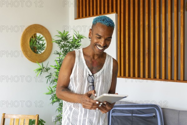 Hispanic LGBTQIA+ man with blue hair happily prepares for holidays using a tablet, surrounded by cozy home decor and greenery. Embracing celebration and diversity